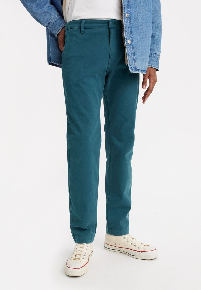 Pantaloni din bumbac teal, cu croială dreaptă, buzunare frontale și închidere cu buton, asortați cu adidași din pânză crem. Geaca din denim este vizibilă.