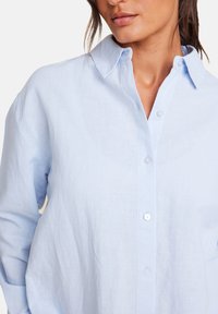 Lyseblå lin skjorte med knaplukning foran og klassisk krave, med en afslappet pasform og glat tekstur. Simple hvide knapper er synlige.
