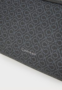 Čierna textúrovaná peňaženka s embosovaným logom Calvin Klein. Obsahuje vzorovaný dizajn prepojených písmen v tmavosivej farbe.