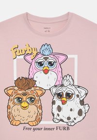 Lyserød t-shirt med tre tegneserie-Furby-figurer med store øjne, i særskilte farver (lyserød, brun, hvid) og teksten "Frigør din indre FURB."