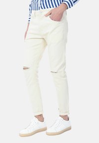 Jeans skinny beige chiaro da uomo con un design strappato e buchi sulle ginocchia, realizzati in cotone con una texture liscia, abbinati a sneakers bianche.
