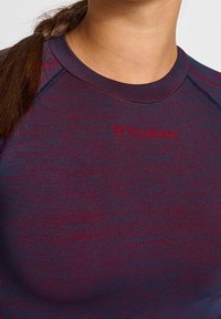 Lähikuva henkilöstä, joka käyttää maroonin ja tummansinisen s�ävyistä tekstuurilla varustettua urheilupaitaa, jossa on "hummel"-logo rinnassa ja ruskea palmikkohiukset olkapäällä.