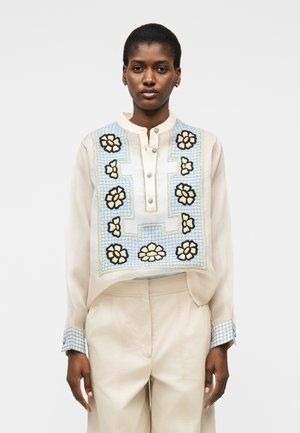 Γυναίκα που φοράει κρεμ μπλούζα με μπλε τετραγωνισμένο σχέδιο και κίτρινη κεντητική με λουλούδια στο εμπρός μέρος και στα μανίκια, συνδυασμένη με ψηλόμεσο κρεμ παντελόνι.