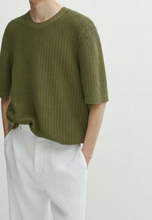 Homme portant un pull en maille à manches courtes texturé vert olive et un pantalon blanc ample, les mains dans les poches, sur un fond uni.