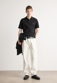 Sort polo shirt med en lille logo, hvide brede bukser og sorte sko. En sort jakke er lagt over den ene arm. Enkel, ren design.