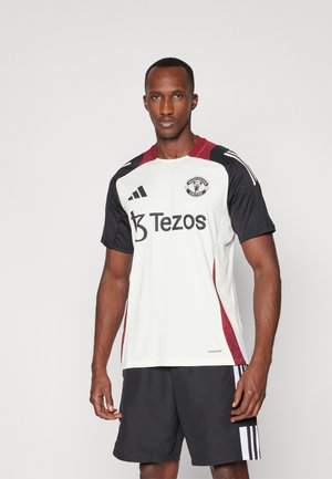 Witte voetbalshirt met korte mouwen, zwarte mouwen, rode zijkanten, met het logo van Manchester United en "Tezos"-branding.