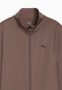 Puma zip-up jakke i blødt brunt stof. Høj krave, fuld lynlås og sort logo på venstre bryst. Glat tekstur uden synlige mønstre.