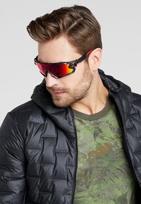 Occhiali da sole con lenti rosse e montatura nera, indossati con un giubbotto nero imbottito e una maglietta verde con motivo camouflage sotto.