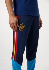 Jūras spēku sporta bikses ar sarkaniem, dzelteniem un atkal sarkaniem sānu svītrainījumiem; rotā tās dzeltena izšūta Spānijas emblēma ar zvaigzni virsū.