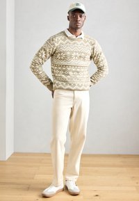 Maglione beige con motivo e disegni geometrici bianchi, abbinato a pantaloni beige e sneakers bianche. Il modello indossa un cappellino chiaro.