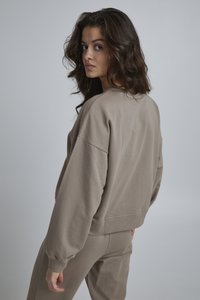 Felpa marrone con spalle scese e vestibilità rilassata, realizzata in tessuto testurizzato. Modello visto da dietro, abbinandola a pantaloni coordinati.