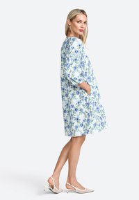 Blumenkleid in Blau- und Grüntönen, mit Dreiviertelärmeln, seitlichen Taschen und lockerer Passform. Kombiniert mit beigen Slingback-Schuhen.