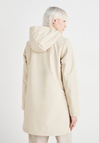 Veste imperméable beige avec capuche, texture lisse, design longueur genou et style minimaliste. Vue arrière montrant des lignes et des coutures épurées.
