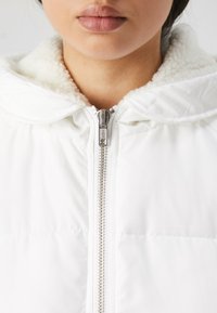 Veste blanche avec une doublure douce et pelucheuse dans la capuche, dotée d'une fermeture éclair argentée et d'un motif matelassé sur le corps.