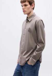 Ανοιχτό καφέ πουκάμισο με κουμπιά και γιακά, με ένα διακριτικό κεντημένο λογότυπο στο στήθος και μακριά μανίκια, σε συνδυασμό με μπλε τζιν.