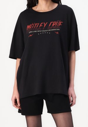 Personne portant un T-shirt noir surdimensionné avec le texte rouge « Mötley Crüe » et un short noir, debout devant un fond blanc.