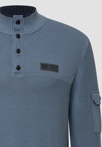 Niebieski sweter z teksturowanego dzianiny z czterema czarnymi guzikami, prążkowanym kołnierzem, małą naszywką z logo na klatce piersiowej oraz kieszonką z klapką na lewym rękawie.