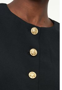 Κοντινό πλάνο μαύρου ενδύματος με τρία περίτεχνα χρυσά κουμπιά κατά μήκος του μπροστινού μέρους κοντά στον γιακά σε σκούρο δέρμα.