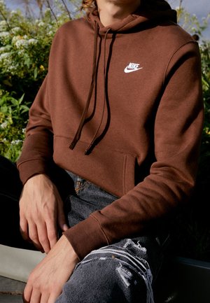 Osoba ubrana w brązową bluzę z kapturem Nike i podarte czarne dżinsy, siedząca na zewnątrz, w tle zieleń.