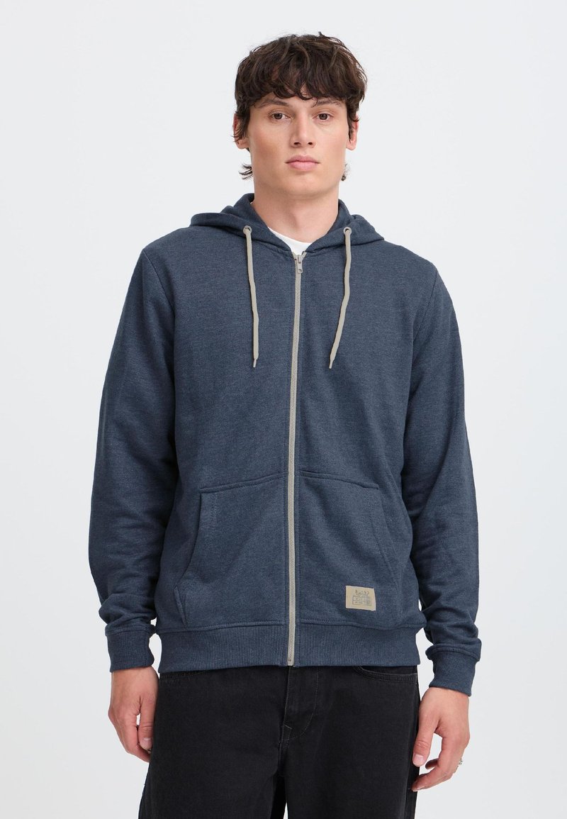Giovane uomo con felpa con cappuccio blu scuro con zip, tasche frontali e coulisse, in piedi davanti a uno sfondo chiaro e uniforme.