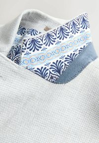 Detailný záber na golier svetlosivej textúrovanej košele s preloženým vnútorným lemovaním s modrým, bielym a oranžovým kvetinovým vzorom.