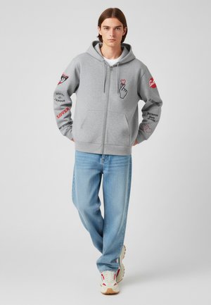 Felpa grigia con zip, dotata di tasca frontale, grafica a mano e testo stampato in rosso e nero sulle maniche. Abbinata a jeans azzurri e sneakers.