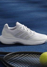 White athletic shoe with three stripes on blue tennis court next to black tennis racket and yellow tennis ball.