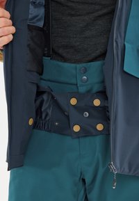 Navy waterproof jacket interior featuring a dark gray fabric, with a teal waistband and bronze snap buttons, and an elasticized waistband for fit.