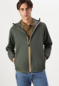 Chaqueta ligera de color verde oliva con cremallera frontal que presenta una franja multicolor, capucha ajustable y puños elásticos. Llevada sobre un polo marrón.