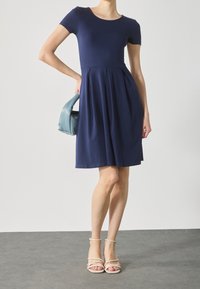 Navy blue short-sleeve dress with a rounded neckline, fitted bodice, and flared skirt; paired with a light blue handbag and neutral sandals.