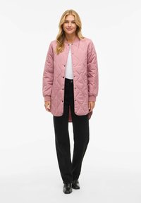 Svetlo roza prešita dolga jakna z okroglim ovratnikom, pritisnimi gumbi in rebrastimi manšetami, kombinirana s črnimi hlačami in gležnjarji.