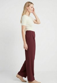 Femme debout de profil, portant un haut crème à manches courtes, un pantalon bordeaux taille haute et des ballerines beiges sur un fond uni.