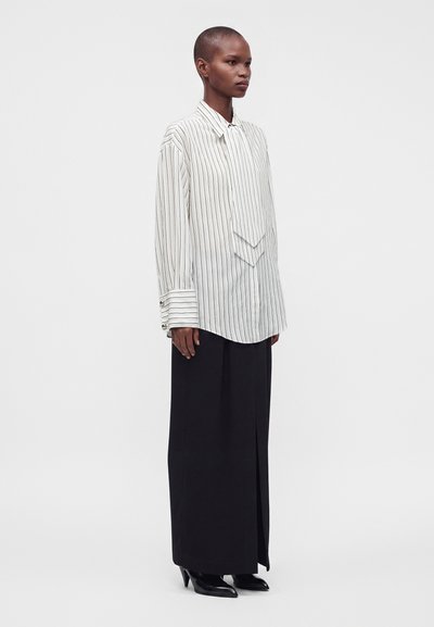 Personne portant une chemise blanche à fines rayures à manches longues avec des poignets surdimensionnés et un pantalon noir à jambes larges avec des chaussures noires à talons, debout de côté.
