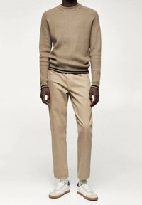 Beige Ripppullover mit kontrastierender schwarzer Streife an den Bündchen, kombiniert mit hellbeigen Hosen und weißen Sneakers mit braunem Profil.