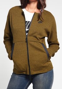 Chaqueta acolchada verde oliva con cierre, con un acabado texturizado, detalles negros en los bolsillos y un pequeño logo en el pecho. Presenta un corte relajado.