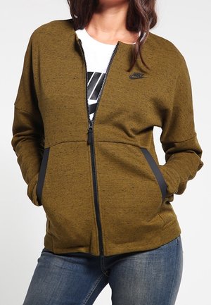 Veste zippée vert olive avec une finition texturée, accents noirs sur les poches et un petit logo sur la poitrine. Présente une coupe décontractée.