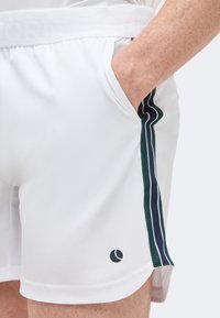 Χέρι στην τσέπη λευκών αθλητικών σορτς με πράσινη και ναυτικό ριγέ πλαϊνή λωρίδα και μικρό μαύρο κυκλικό λογότυπο κοντά στο στρίφωμα.