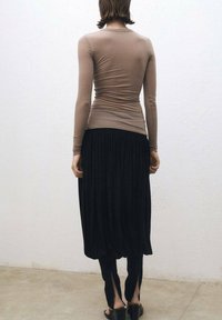 Beige Langarmshirt, kombiniert mit einem schwarzen, plissierten Rock und schmal zulaufenden Hosen. Glatte Textur mit dezenten Drapierungen. Sandalen mit dicken Sohlen.