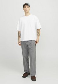 White short-sleeve t-shirt with a loose fit, paired with gray straight-leg jeans and brown shoes. The background is neutral and light.