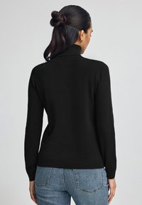 Kobieta o ciemnych włosach związanych w półkucyk, ubrana w czarny golf i niebieskie dżinsy, pokazana od tyłu na jednolitym tle.