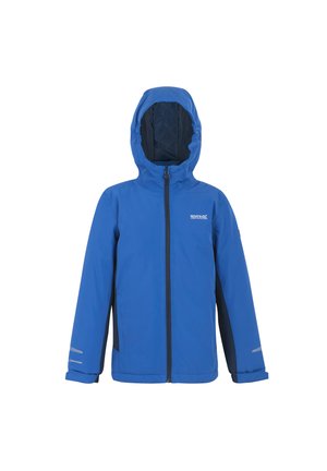 Jongen glimlachend met blauwe waterdichte jas, grijze trui met rits, marineblauwe broek en zwart-groene sneakers, staand tegen een lichte achtergrond.