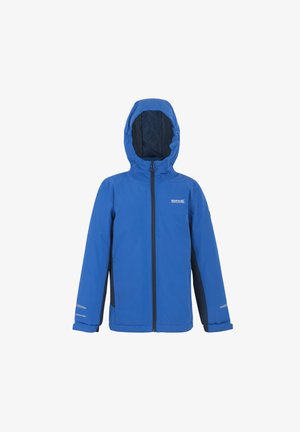 Niño sonriendo con chaqueta impermeable azul, suéter gris con cremallera, pantalones azul marino y zapatillas negras y verdes, de pie contra un fondo claro.