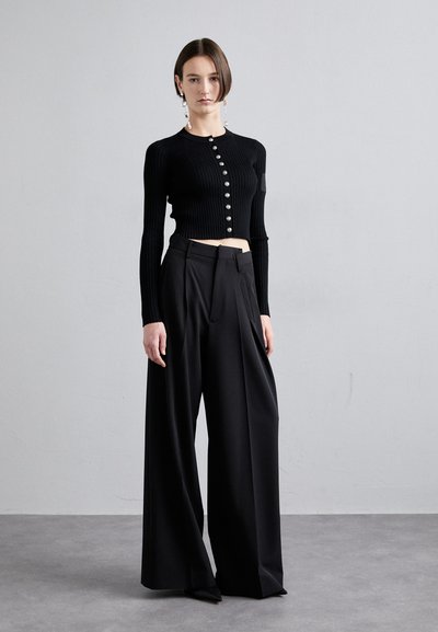 Zwart geribde crop top met knoopsluiting, gecombineerd met zwarte high-waisted wijde broek. Model staat tegen een neutrale achtergrond.