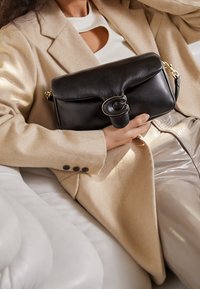 Czarna skórzana torebka z okrągłym złotym zapięciem, o miękkim, strukturalnym wyglądzie. Torebkę trzyma osoba ubrana w beżowy płaszcz.