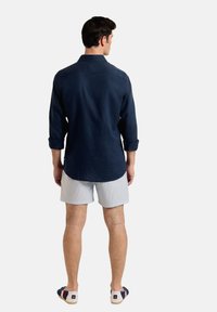 Camisa de manga larga azul marina con puños abotonados, llevada sobre pantalones cortos a rayas ligeras. Sandalias informales completan el atuendo. Fondo liso.
