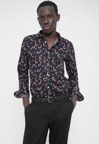 Schwarzes Hemd zum Zumachen mit Zuckerstangen- und Blumenmuster. Lange Ärmel mit abgerundetem Manschettenabschluss, aus leichtem Stoff. Kombiniert mit schwarzer Hose.