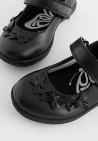 Czarne skórzane buty typu Mary Jane z ozdobnymi akcentami w kształcie motyla, dwiema regulowanymi klamerkami i teksturowaną podeszwą. Widoczna miękka podszewka.