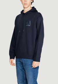 Felpa blu navy con tasca a marsupio, cappuccio con cordini e toppa con logo sul petto. Polsini e orlo a costine per una vestibilità aderente.