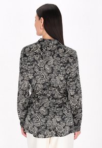 Maglietta nera a maniche lunghe con motivo paisley bianco, texture morbida, colletto e orlo dritto, indossata sbottonata.