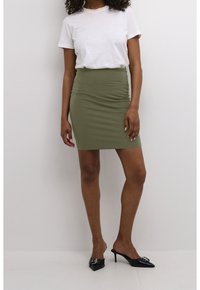 Femme portant une chemise blanche à manches courtes, une jupe crayon taille haute olive, et des mules à talons aiguilles noires sur fond blanc.